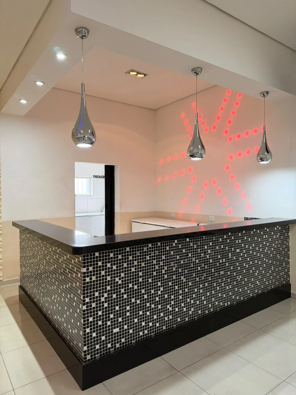 Bar em L revestido em pastilhas preto e branco, com pendentes cromados e luz de LED na parede